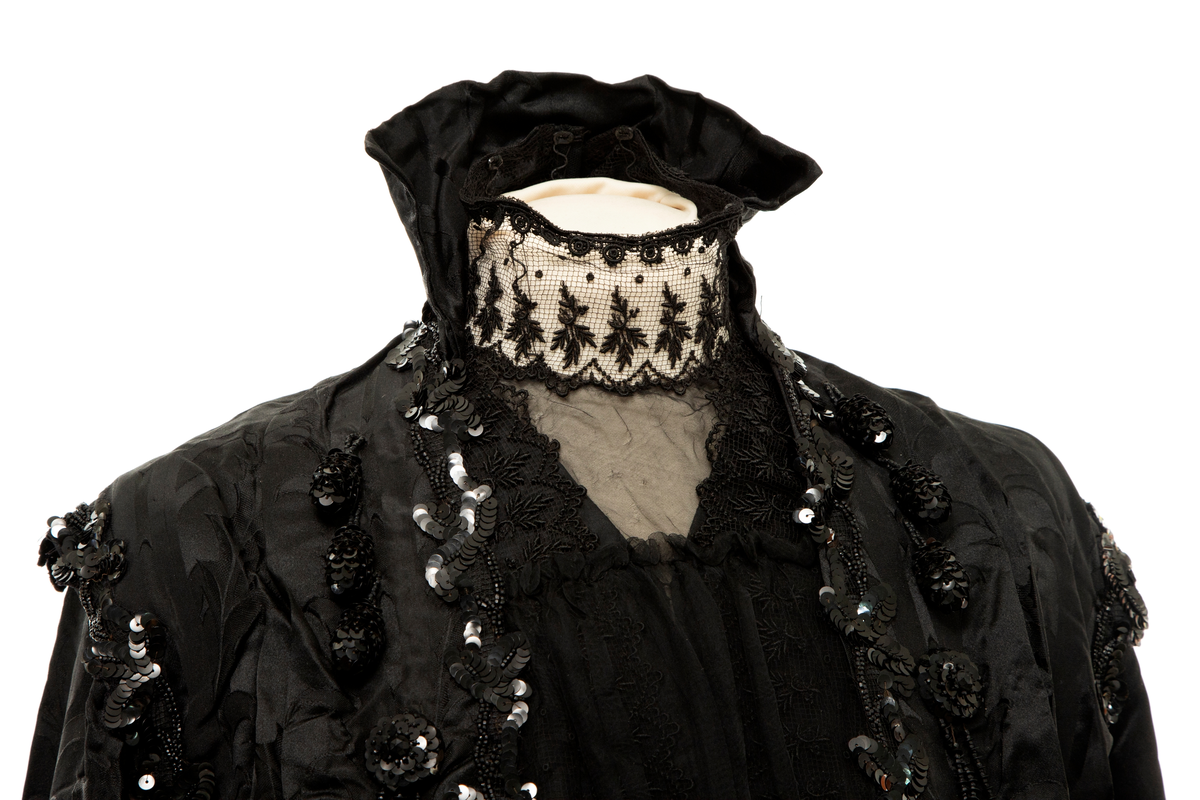 Sortfarget kvinnedrakt i tre deler:

1) Kjoleliv, midjelangt, med frontlukning, lange, trange ermer, delvis løs vest over bysten, chiffon "duebryst" og blonde ståkrage innenfor en Maria Stuart-krage. Mye dekorativ bruk av paljetter og steinkullsperler i broderier og possementeri. Fôret.

2) Skjørt, utskrådd og kilesydd, forsiktig rynket til smal linning, noe mer rynket midt bak, på hver side av lukningen. Lukket med hekter. Ufôret.

3) Belte, drapert og ufôret. Splittet i to i hver ende. Endestykkene lukkes med henholdsvis én og to trykknapper skjult av hver sin sløyfe. Sydd for hånd.