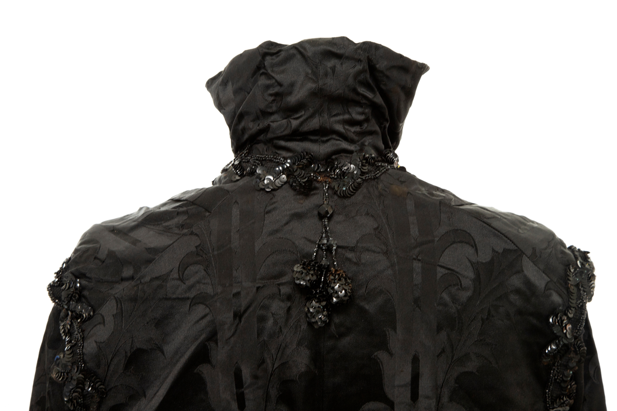 Sortfarget kvinnedrakt i tre deler:

1) Kjoleliv, midjelangt, med frontlukning, lange, trange ermer, delvis løs vest over bysten, chiffon "duebryst" og blonde ståkrage innenfor en Maria Stuart-krage. Mye dekorativ bruk av paljetter og steinkullsperler i broderier og possementeri. Fôret.

2) Skjørt, utskrådd og kilesydd, forsiktig rynket til smal linning, noe mer rynket midt bak, på hver side av lukningen. Lukket med hekter. Ufôret.

3) Belte, drapert og ufôret. Splittet i to i hver ende. Endestykkene lukkes med henholdsvis én og to trykknapper skjult av hver sin sløyfe. Sydd for hånd.