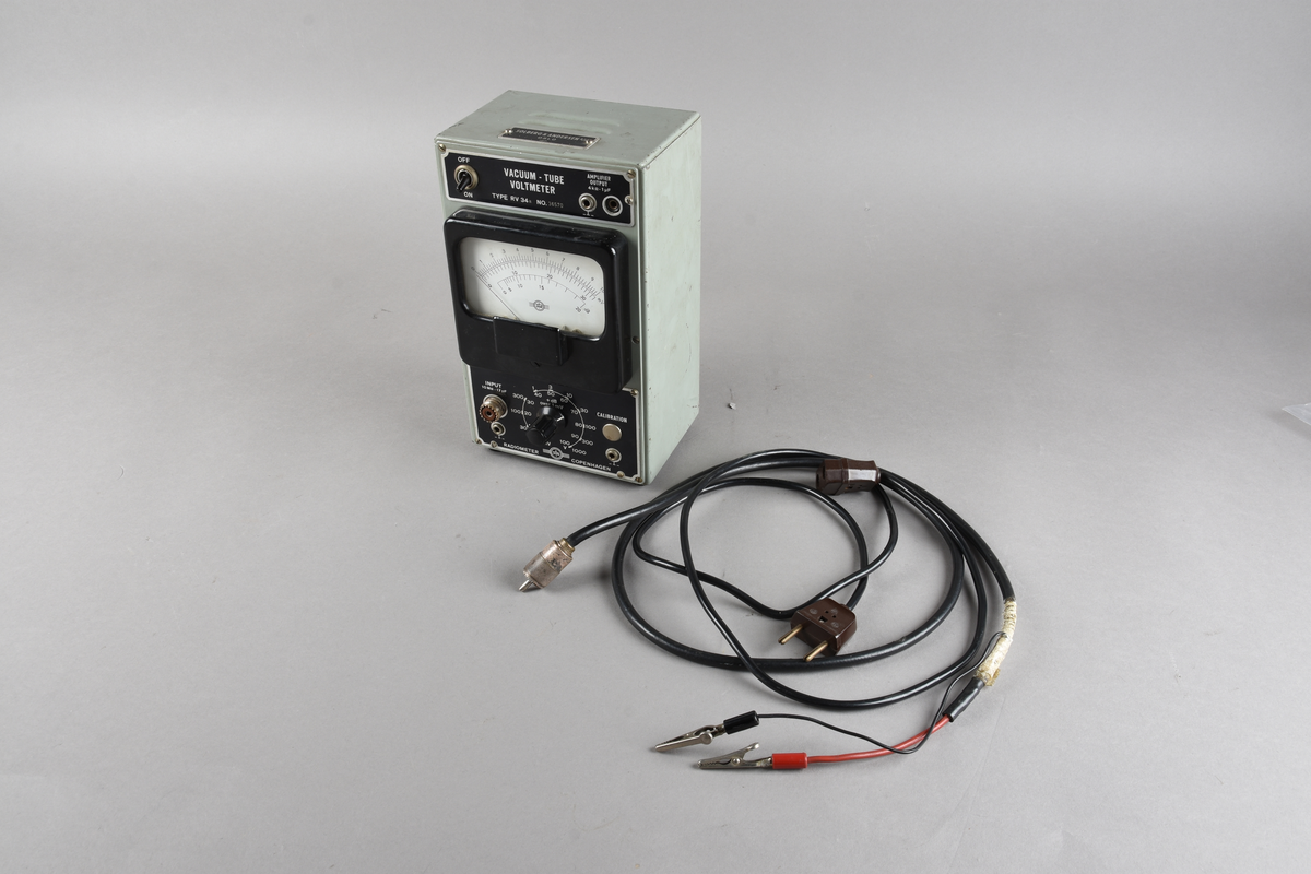 Voltmeter i grågrønn rektangulær utforming, men sorte knapper og hvitt vindu i front. Håndtak og knotter for stående stilling i underkant. To løse ledninger følger med.