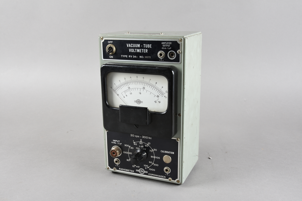 Voltmeter i grågrønn rektangulær utforming, men sorte knapper og hvitt vindu i front. Håndtak og knotter for stående stilling i underkant. To løse ledninger følger med.