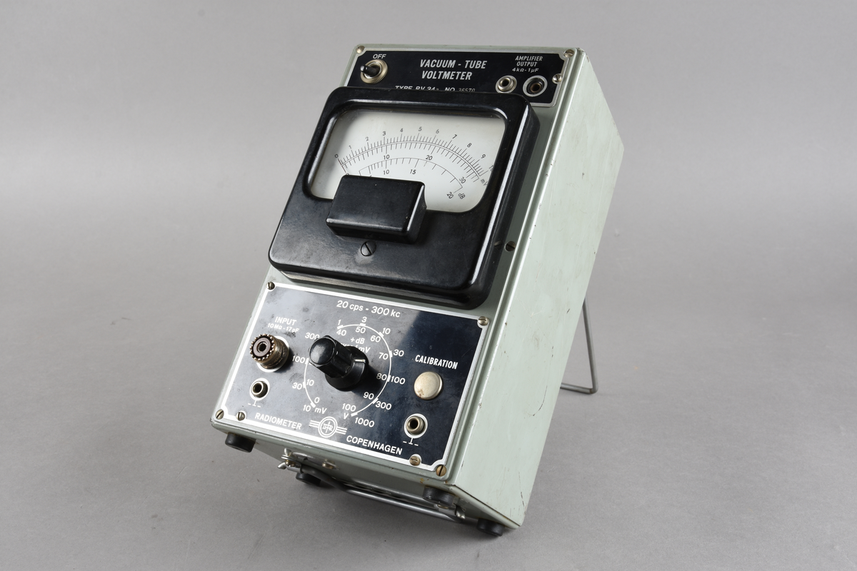 Voltmeter i grågrønn rektangulær utforming, men sorte knapper og hvitt vindu i front. Håndtak og knotter for stående stilling i underkant. To løse ledninger følger med.