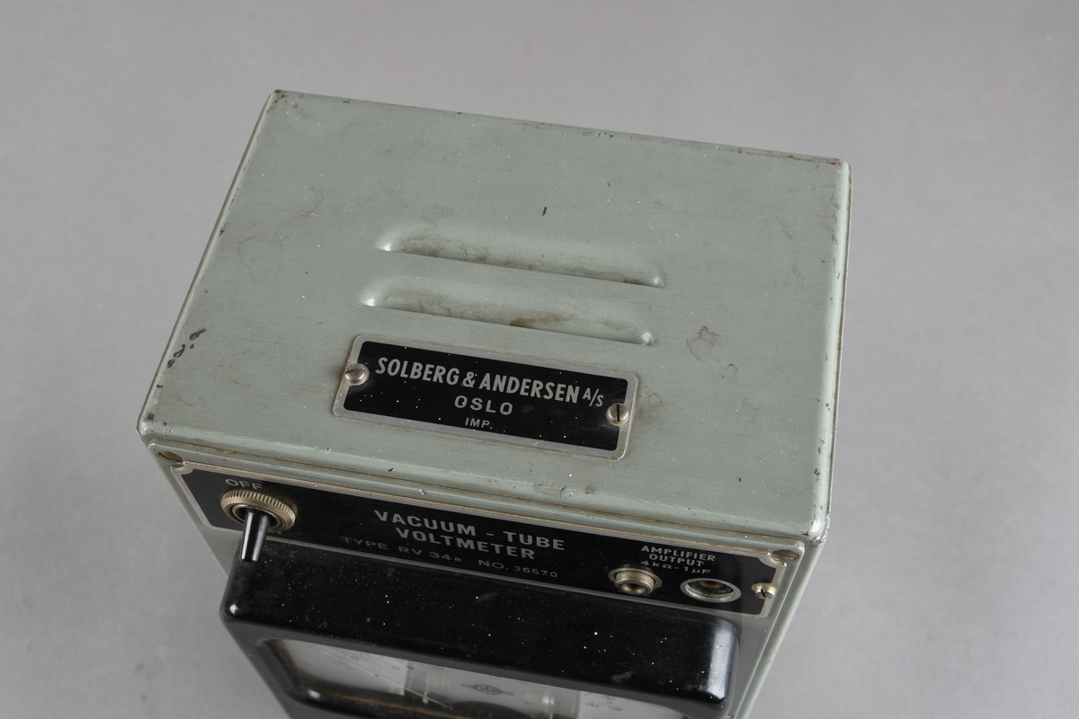 Voltmeter i grågrønn rektangulær utforming, men sorte knapper og hvitt vindu i front. Håndtak og knotter for stående stilling i underkant. To løse ledninger følger med.