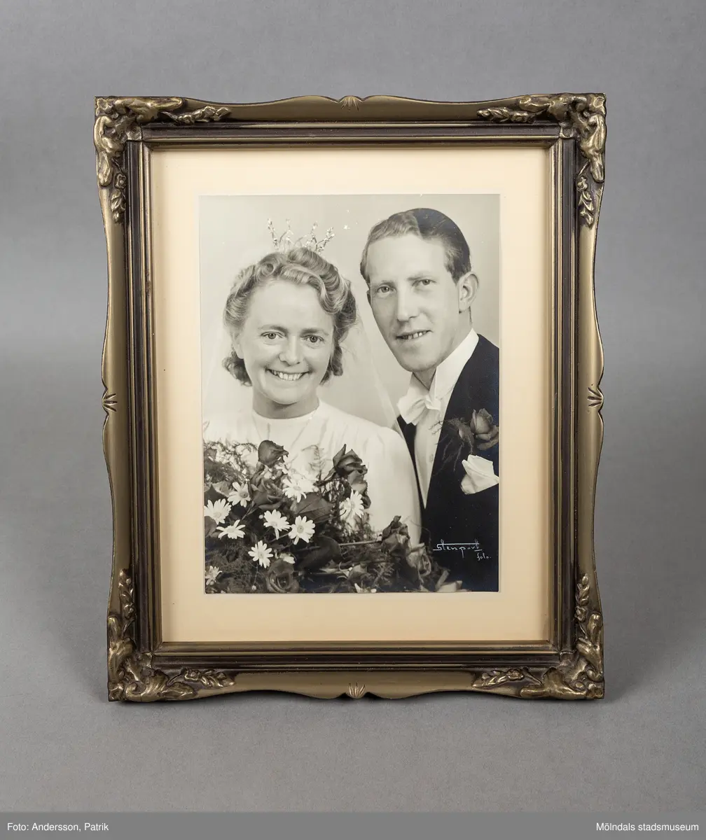Ett svartvitt inramat bröllopsfoto. Bruden som är till vänster har en bröllopskrona på huvudet. Hon håller en stor blomsterbukett bestående av vita Margeriter och rosor. Bruden bär en vit rundhalsad klänning. Brudgummen har stärkt vit krage, svart frack med vit näsduk och en ros i frackfickan. Fotografiets ram är av trä och gråfärgad. Ramen har blommönster i hörnen och reliefmönster på sidorna.
Porträttet föreställer Karin Hanssons föräldrar. Karin har lämnat flera andra föremål till vår samling.