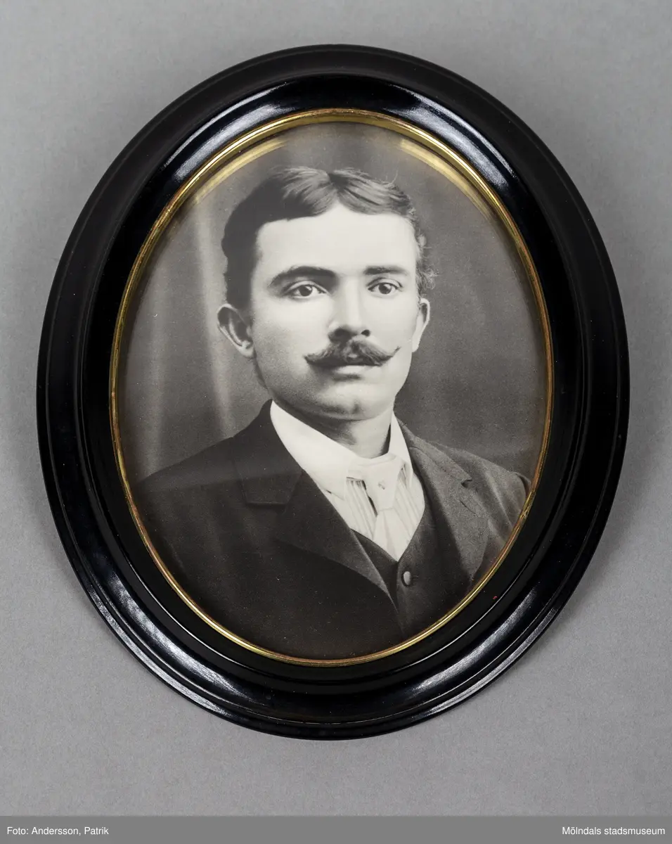 Ett porträtt inramat i en oval ram. Ramen är svart med en guldkant. Porträttet föreställer en ung man med mustasch. Mannen är klädd i vit skjorta, väst och kavaj. Han bär också en vit slips.