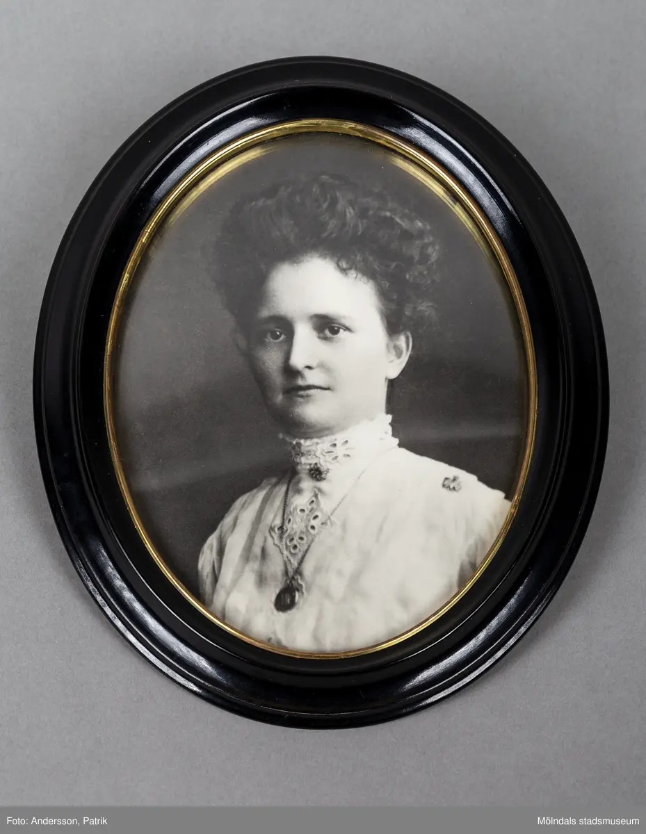 Ett porträtt inramat i en oval ram. Ramen är svart med guldkant. Kvinnan är klädd i en vit blus med hög krage och hålmönster. Över blusen bär hon ett halsband med ett ovalt smycke. Vid kragen finns en brosch fäst. Kvinnan heter Nora Krantz och är mormor till Karin Hansson.