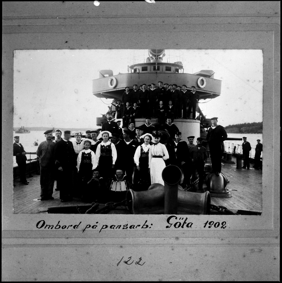 Denna grupporträtt föreställer manskap och besökare ombord på pansarbåten Göta 1902. I förgrunden sitter två sjömän med musikinstrument. Bakom de har några kvinnor i traditionella dräkter tagit uppställning för en gruppfoto. I bakgrunden har manskapet samlats ovanpå kupolen av kanontornet.