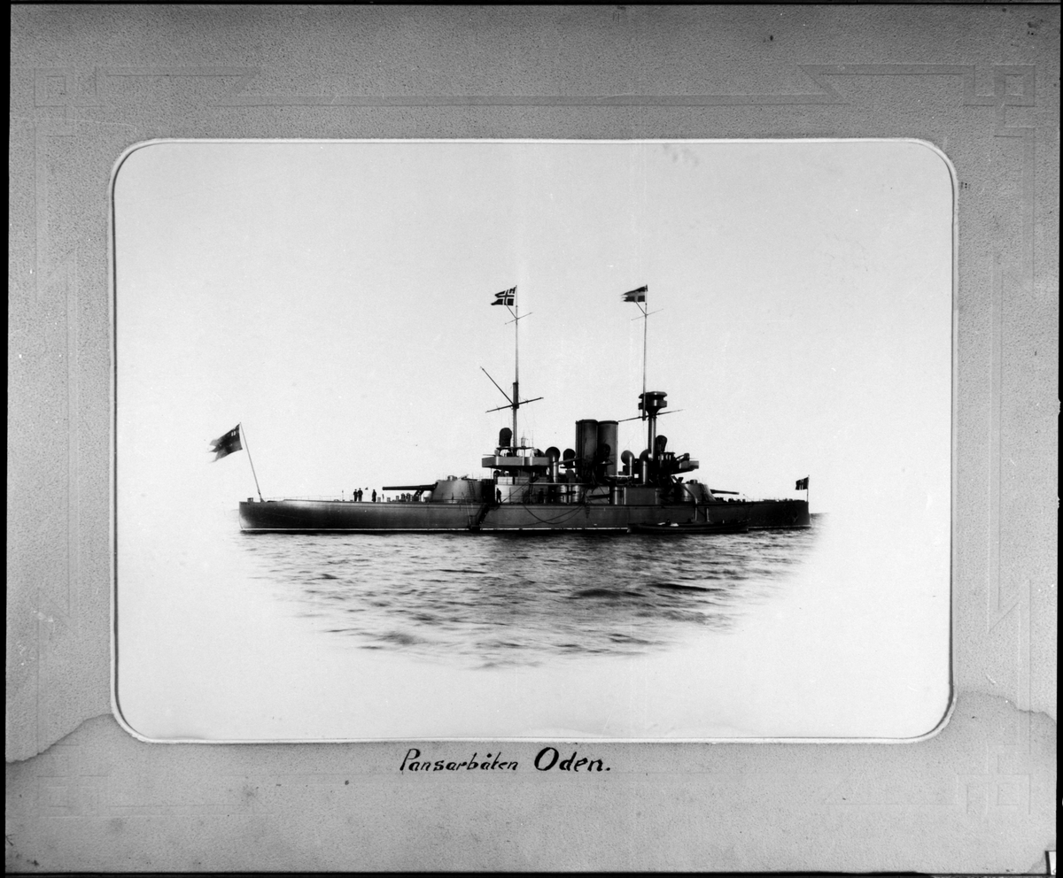 Fartygsporträtt som visar pansarbåten Oden till ankars. Fartyget syns från babordssidan där ett mindre ångbåt ligger förtöjt. Flera repstege leder upp till däcket. På akterdäcket står tre men som verka samtala.