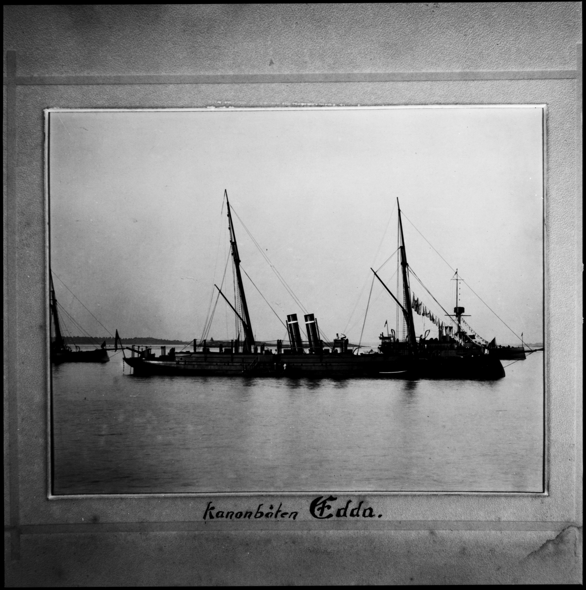 Fartygsporträtt som visar kanonbåten Edda till ankars. Fartyget syns från babordssidan där två skeppsbåtar ligger förtöjt. I bakgrunden syns ett skärgårdslandskap och ytterligare två kanonbåtar.