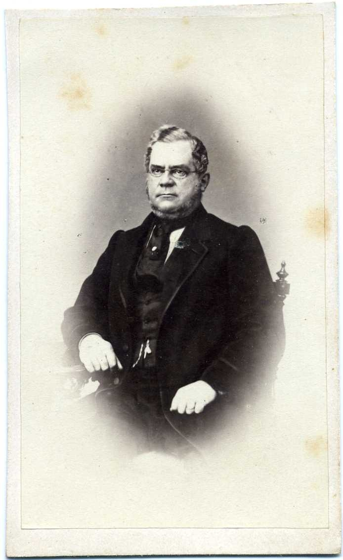 Porträtt av en riksdagsman med glasögon, sittande på en stol vid ett bord.