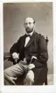 Porträtt av en man sittande på en stol vid ett bord med en spatserkäpp i vänster hand. Troligen riksdagsman, eventuellt G Hultman. Autograf på bildens baksida.