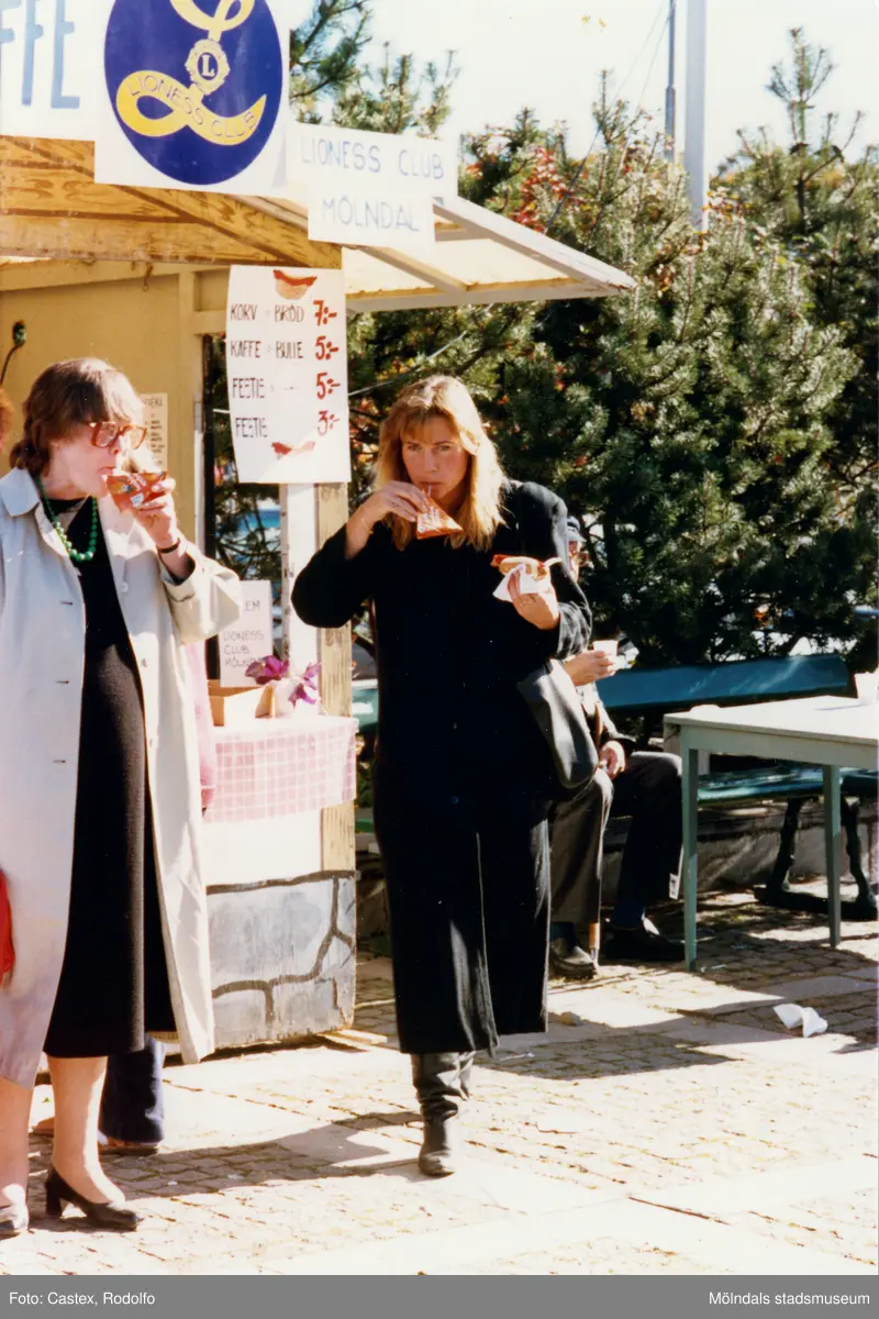 Lions Club anordnar marknad på Stadshusplatsen i Mölndal, tidigt 1980-tal. Två kvinnor äter korv med bröd och dricker Sunkist Apelsin till.