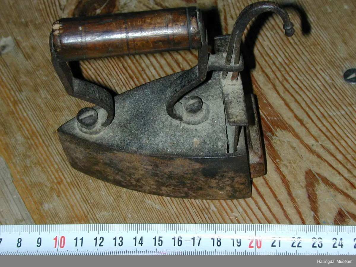 Tre loddrette sider, dei to langsidene noko krumma, stuttsida rett med opning til bolt. Over- og undersida flat. På oversida eit rektangulært hol på den bakre delen. I holet ei jarnplate med stang med bøygd krok øvst midt på. Denne jarnplate tener som lem for bolten som er fastrusta inni strykejarnet. Sylindrisk dreia handtak.