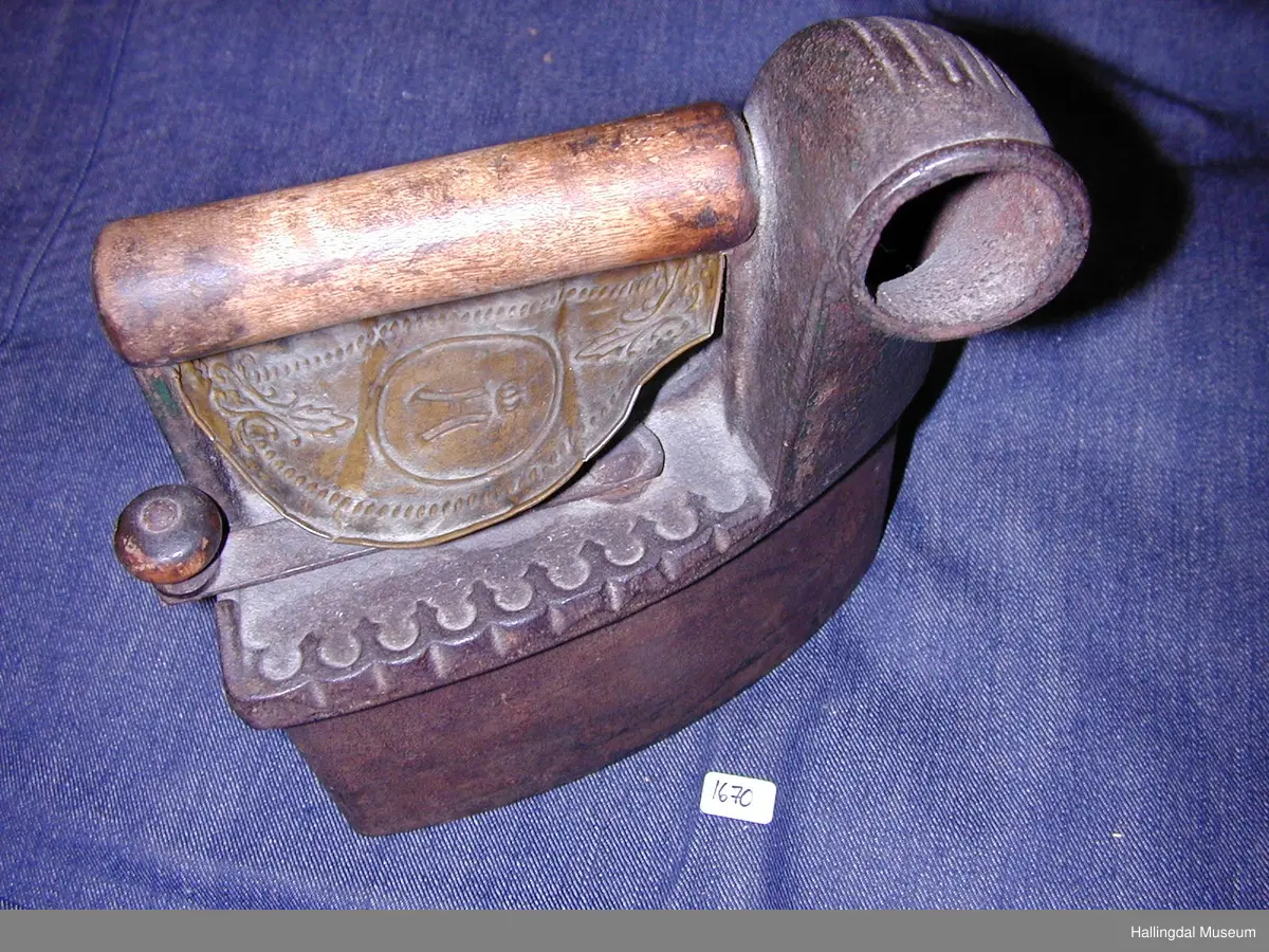 Strykejarn støypt i jarn og med dreia hank. Oversida er eit lok for å kunne putte glør i strykejarnet. Under handtaket ei tunn messingplate med gravert ornamentikk og bokstaven H med krone over i midten.