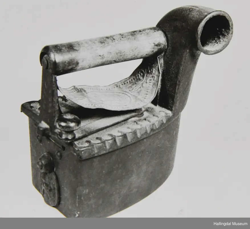 Strykejarn støypt i jarn og med dreia hank. Oversida er eit lok for å kunne putte glør i strykejarnet. Under handtaket ei tunn messingplate med gravert ornamentikk og bokstaven H med krone over i midten.