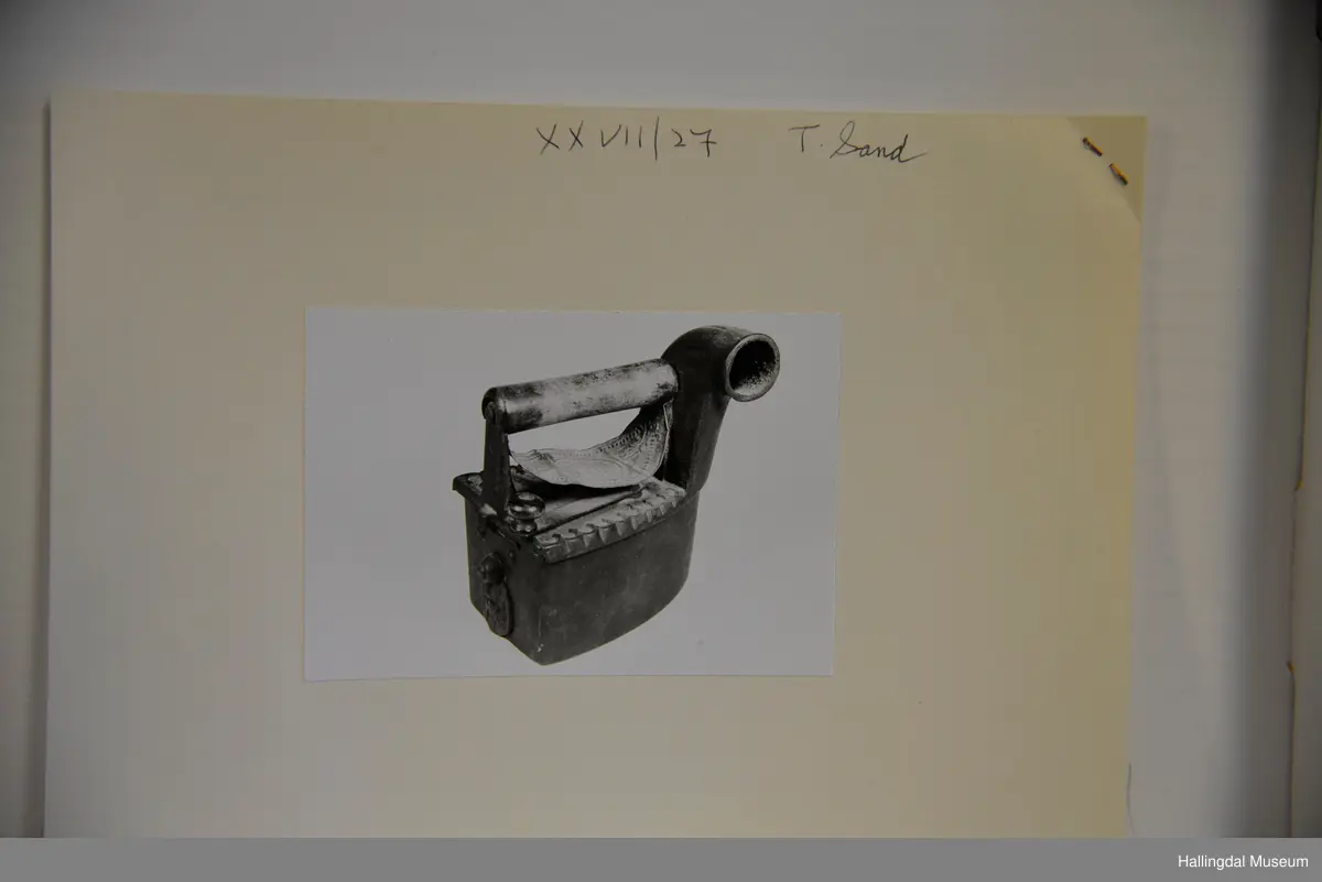 Strykejarn støypt i jarn og med dreia hank. Oversida er eit lok for å kunne putte glør i strykejarnet. Under handtaket ei tunn messingplate med gravert ornamentikk og bokstaven H med krone over i midten.