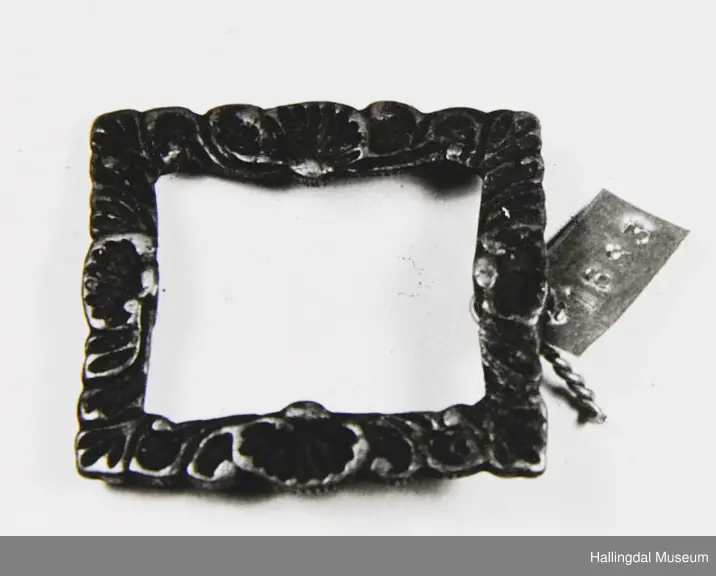 Del av spenne i messing (?). Rektangulær ramme, støypt blomsterornamentikkk, glatt på baksida. Stanga er borte.