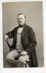 Porträtt av en riksdagsman sittande på en stol. Han bär ljusa smårutiga byxor och väst. Autograf på bildens baksida.