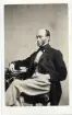 Porträtt av en riksdagsman sittande på en stol vid ett bord, med armen vilande på böcker. Han bär ljusa byxor och väst. Autograf på bildens baksida.