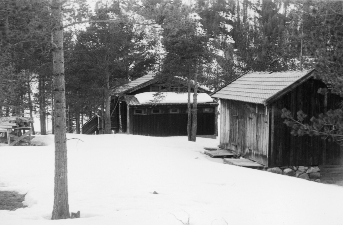 Fra «Maribu», ei hytte ved Olstappen i Vinstravassdraget i Nord-Fron i Oppland fylke. Dette fotografiet ble tatt på seinvinteren i 1960. Til høyre i forgrunnen ser vi en uthusbygning, utført i bordkledd bindingsverk med sementsteintekket saltak. Det er nærliggende å anta at dette huset må ha inneholdt utedo og vedskåle. Sentralt i bildet, men mer i bakgrunnen, ser vi sjølve hytta, som var en laftekonstruksjon, men med en tilbygd bindingsverksgang med pulttak på den gavlveggen som vendte mot fotografen. Hytta er avbildet fra en annen vinkel på SJF.1990-03212. Bygningene sto i glissen furuskog. Ved venstre bildkant ser vi et stativ som det lå en del gamle tømmerstokker på.