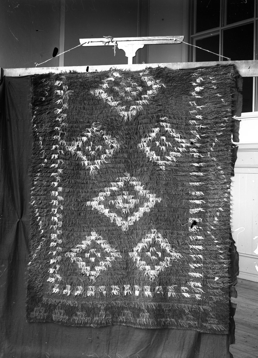 Rya med mönster av romber, ihopsytt av två våder.

Garnprov i kuvert med följande information: Rya, flossa, 130 x 151 cm, 20 rör på 10 cm, varp linne, inslag ullgarn, mönstergarn 2 trådigt ullgarn, Nockans längd utanför knuten 35mm, trådantal i nockan 1, inslag mellan nockraderna 3cm.