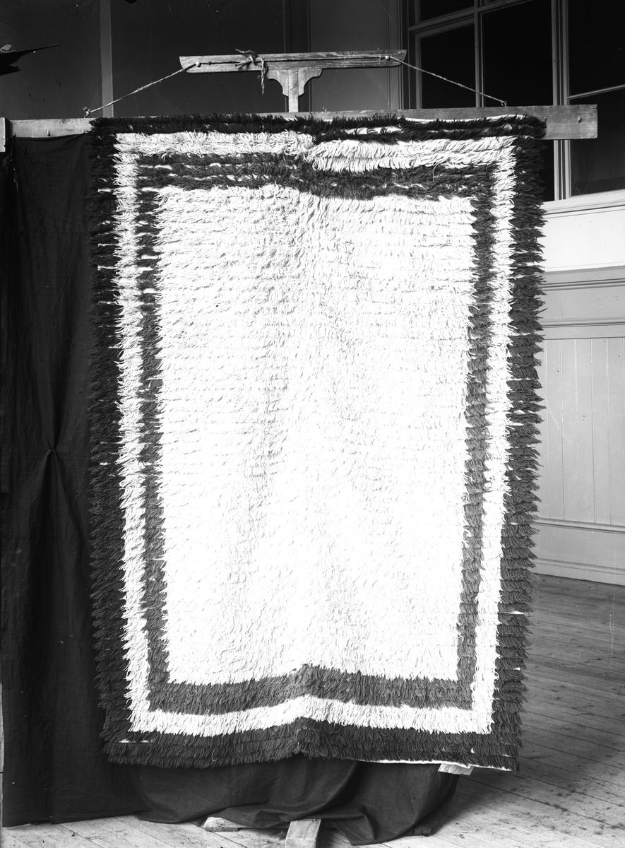 Rya, inramad enfärgad botten.

Garnprov i kuvert med följande information: Rya, flossa, 125 x 183 cm, 40 rör på 10 cm, varp linne, inslag ullgarn, mönstergarn 2 trådigt ullgarn, Nockans längd utanför knuten 32mm, trådantal i nockan 2, inslag mellan nockraderna 28mm.