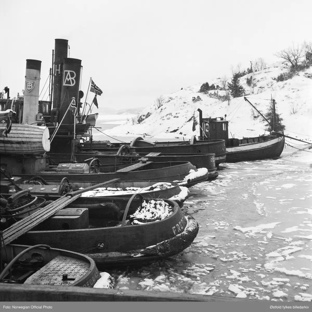 Slepebåtene fra Østfold er vel framme i Sverige etter slepebåtaksjonen 8. februar 1945. 
Fra venstre slepebåtene Per, Fix, Tor,  Oscarsborg, Fram og Marnet.

Tekstseddel limt på bakside av positiv:
NORWEGIAN OFFICIAL PHOTO NO. S. 5570. 
NORSK FLÅTE FØRT TIL SVERIGE.
"Bevepnede menn satte seg torsdag den 8. februar 1945 i besittelse av Fredrikstads hele flåte av slepebåter og dirigerte den til Sverige, ialt 11. Samtidig bergingsbåten "Uredd" som var stasjonert i Moss, dirigert samme vei. Alle disse båter arbeidet for tysk regning med å bugsere fartøyer til ppg fra Fredrikstad,  som er bllitt en stor utskipingshavn for tyskerne. 
Det store kuppet foregikk på den måten at vepnede folk gikk ombord på båtene og overmannet mannskapene. Tre slepebåter som ikke hadde dampen oppe, ble senket på havnen. Kuppet var vel organisert. De kaprende mannskaper gikk ombord i de båter som lå ved kai, og disse båtene ble så styrt bort til de andre som i sin tur fikk kapringsmannskaper ombord. Båtenes kapteiner fikk valget mellom å styre etter ordreeller gå under dekk. En av båtene som skulle  i tyske tjeneste den dagen ble  entret kl. 7.30 om morgenen, og den utførte hele dagen sin slepning av tyske skyteskiver. Først da skyteøvelsene var ferdig, stakk den til Sverige. De andre fartøyer foretok ferden til Sverige i samlet følge.. 
På rad og rekke ligger båtene ien svensk fjord med norske flagg blafrende fra stengene.
Stockholm 22. 2.45."