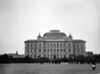 Foto av Växjö Högre Allmänna Läroverk. På skolgården håller man på med att  marschera m.m. Till höger skymtar en flicka. 
I bakgrunden skymtar dåv. borgmästare Bergendahls hus till vänster och till höger ser man bokhandlare Quidings bostad.
