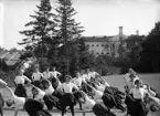 En trupp kvinnliga gymnaster övar i det fria utanför dåv. seminariet, dvs numera Ringsbergsskolan i Växjö.  I bakgrunden syns skolans växthus och dåv. cellfängelset.
