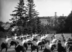 En trupp kvinnliga gymnaster övar i det fria utanför dåv. seminariet, dvs numera Ringsbergsskolan i Växjö.  I bakgrunden syns skolans växthus och  dåv. cellfängelset.