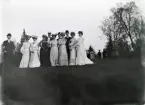 Foto av en samling damer i hatt m.m. tillsammans med två herrar, i någon slags parkanläggning.
Suddigt foto.