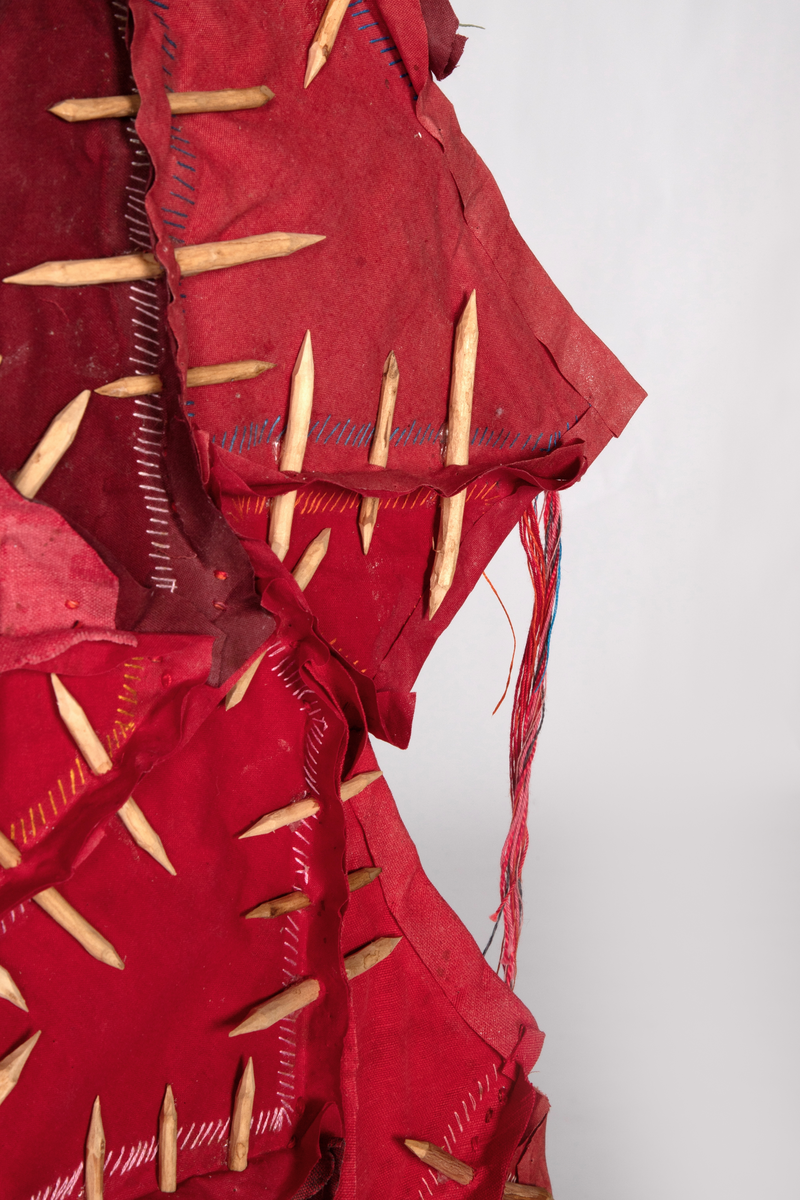 Fritthengende tekstilt romobjekt. Minner om en skinnfell.  Lagd av bekledningstekstiler i bomull, silkefór fra slips, broderi, hudlim, floss i lin og bomull, spikkede bjørkepinner farget med kaffe, oppheng av viklede snorer og y-kvist (suorgi).