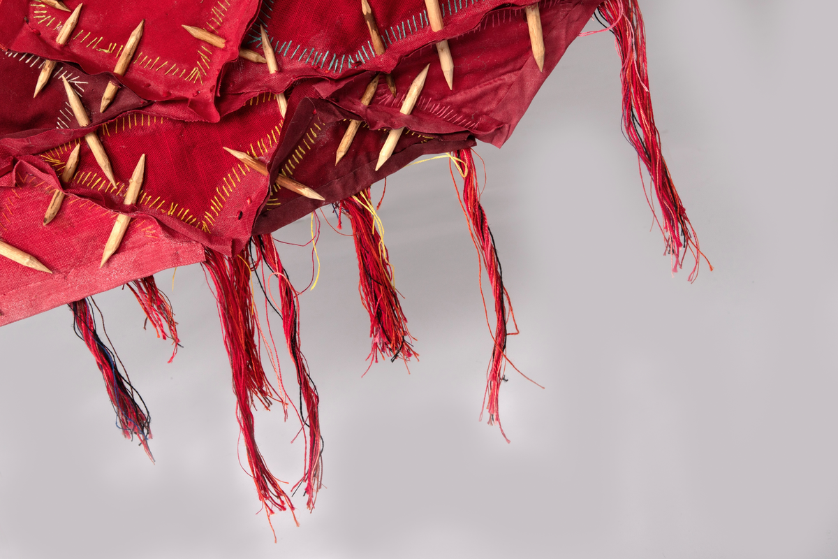 Fritthengende tekstilt romobjekt. Minner om en skinnfell.  Lagd av bekledningstekstiler i bomull, silkefór fra slips, broderi, hudlim, floss i lin og bomull, spikkede bjørkepinner farget med kaffe, oppheng av viklede snorer og y-kvist (suorgi).