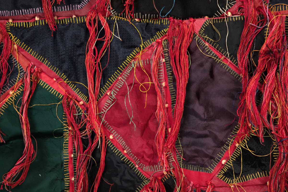 Fritthengende tekstilt romobjekt. Minner om en skinnfell.  Lagd av bekledningstekstiler i bomull, silkefór fra slips, broderi, hudlim, floss i lin og bomull, spikkede bjørkepinner farget med kaffe, oppheng av viklede snorer og y-kvist (suorgi).