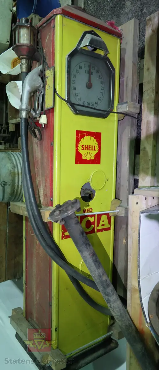 Elektrisk drevet bensinlpumpe med røde sider og gul front. Tekst: SHELL MED ICA. Pumpa har tallskive og viser som går til 100 liter. Kuppel mangler
