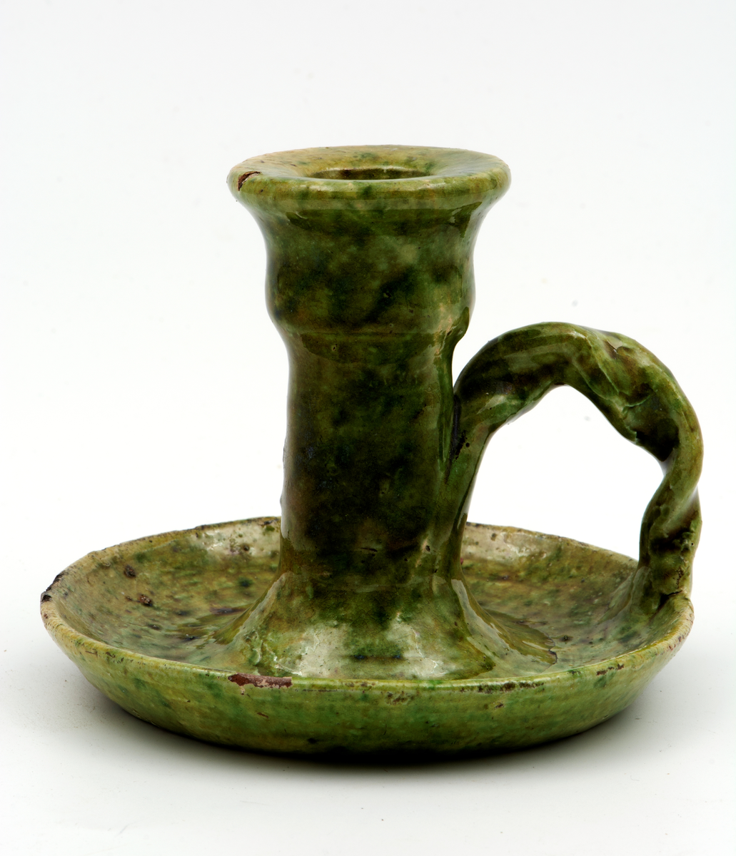 Handdrejad ljusstake av lergods, typ kontorsljusstake. Ljushållare för ett ljus på rund fotplatta med vriden grepe. Ljusgrön bottenlasyr med gröna och bruna sänk. Fotplattan är sprucken tvärs över. På undersidan oläslig text.