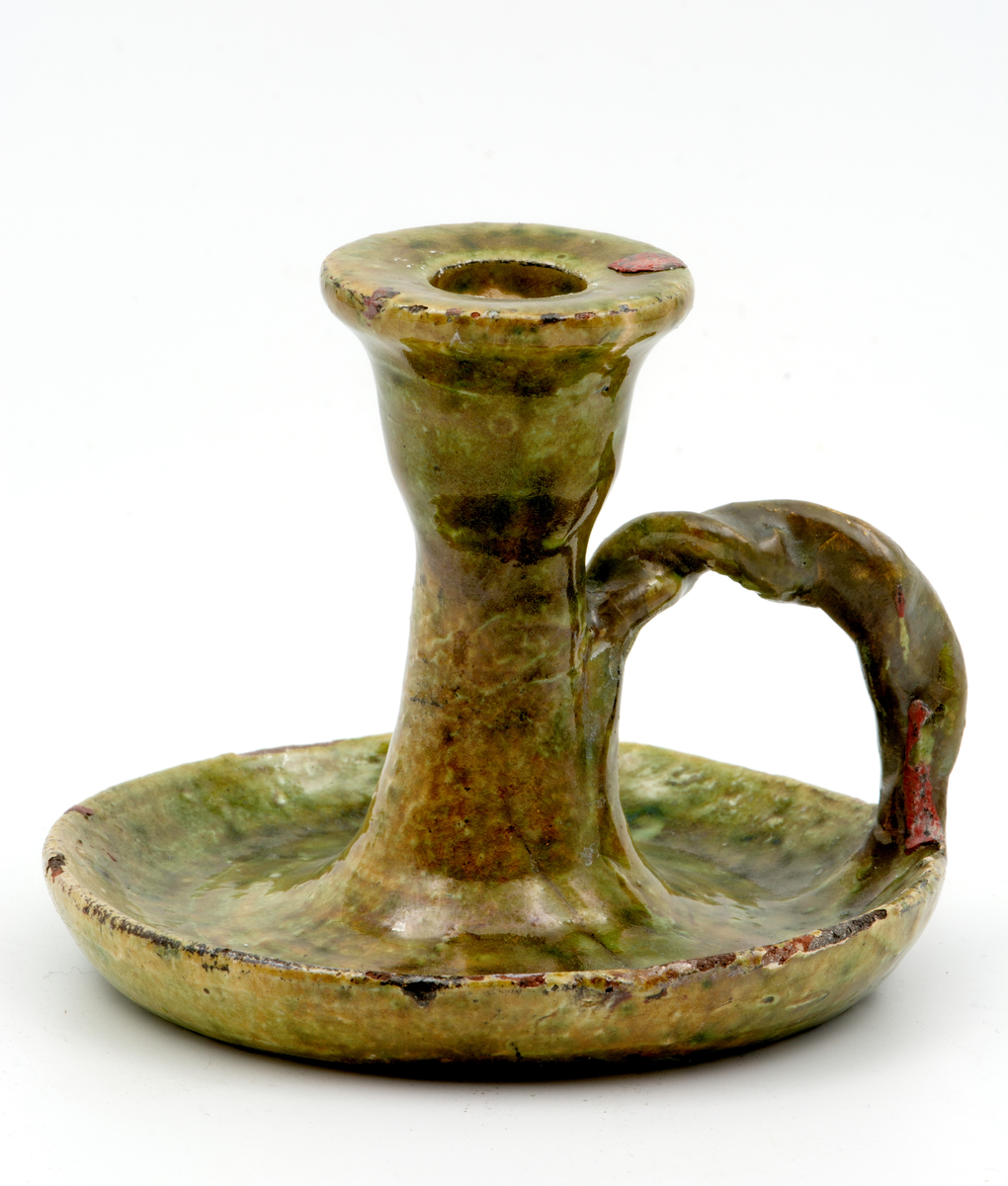 Handdrejad ljusstake av lergods, typ kontorsljusstake. Ljushållare för ett ljus på rund fotplatta med vriden grepe. Ljusgrön bottenlasyr med gröna och bruna sänk. Fotplattan är sprucken tvärs över. På undersidan oläslig text.