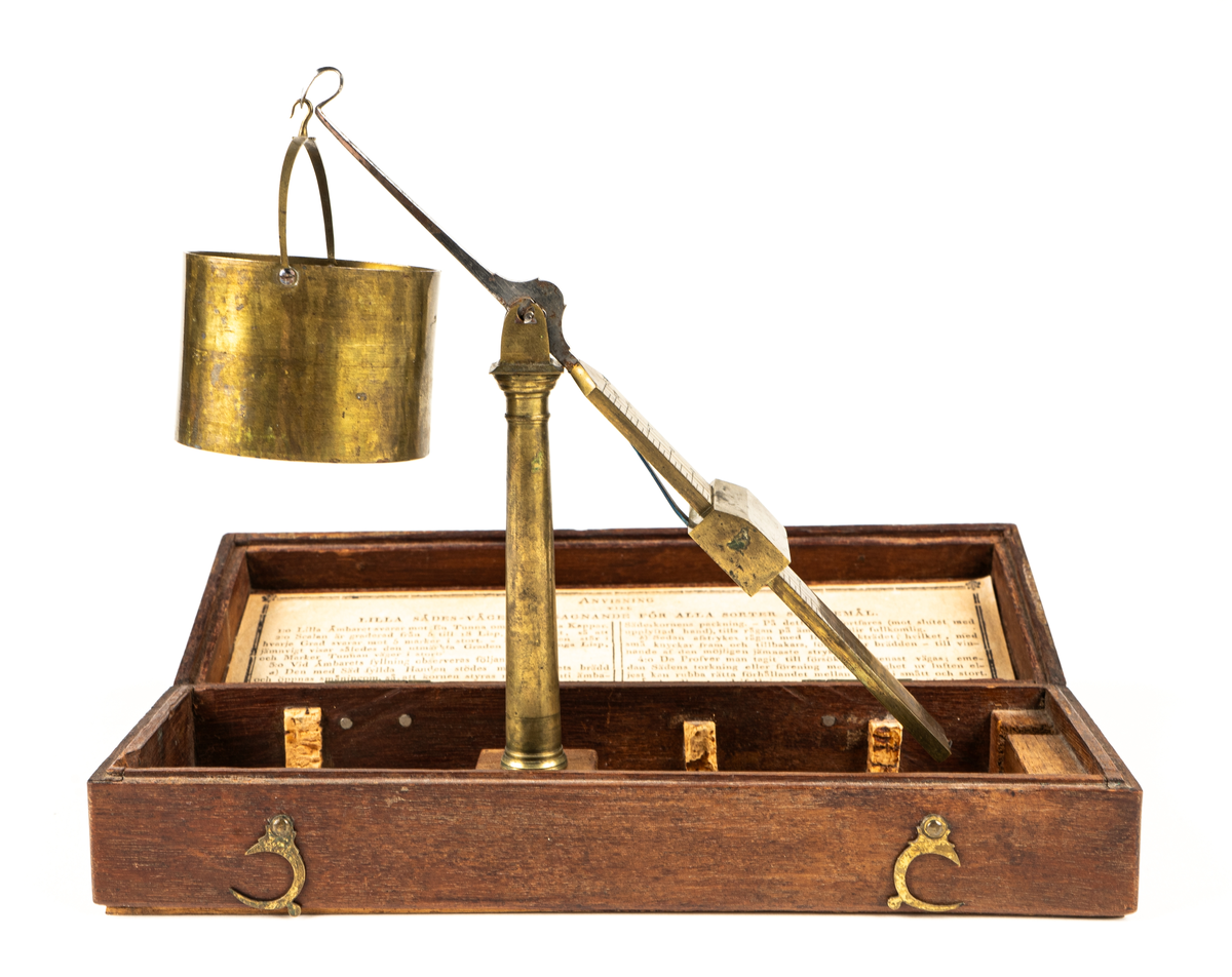 Våg av mässing, för uträkning av spannmålsvikt. Liten provvåg i fodral av trä. Måtten anger fodralets mått.