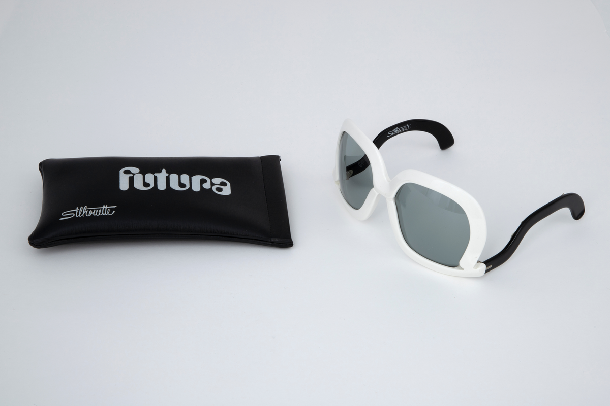 Store solbriller med hvit plastinnfatning og svarte, svungne stenger som er festet nederst på innfatningen. Grønne plastglass. Svart, rektangulært plastetui med åpning på høyre kortside.