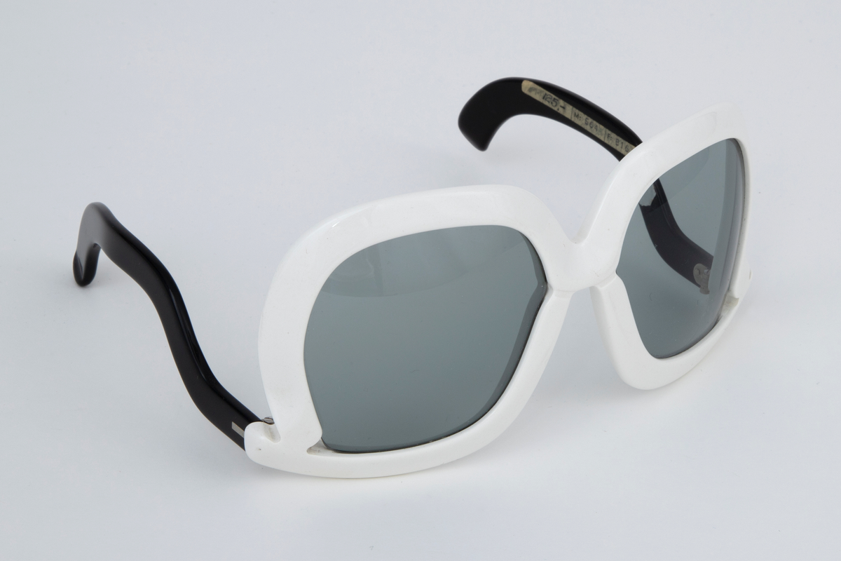 Store solbriller med hvit plastinnfatning og svarte, svungne stenger som er festet nederst på innfatningen. Grønne plastglass. Svart, rektangulært plastetui med åpning på høyre kortside.