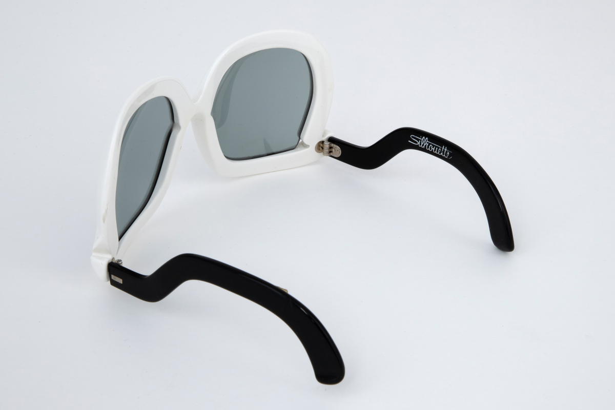 Store solbriller med hvit plastinnfatning og svarte, svungne stenger som er festet nederst på innfatningen. Grønne plastglass. Svart, rektangulært plastetui med åpning på høyre kortside.