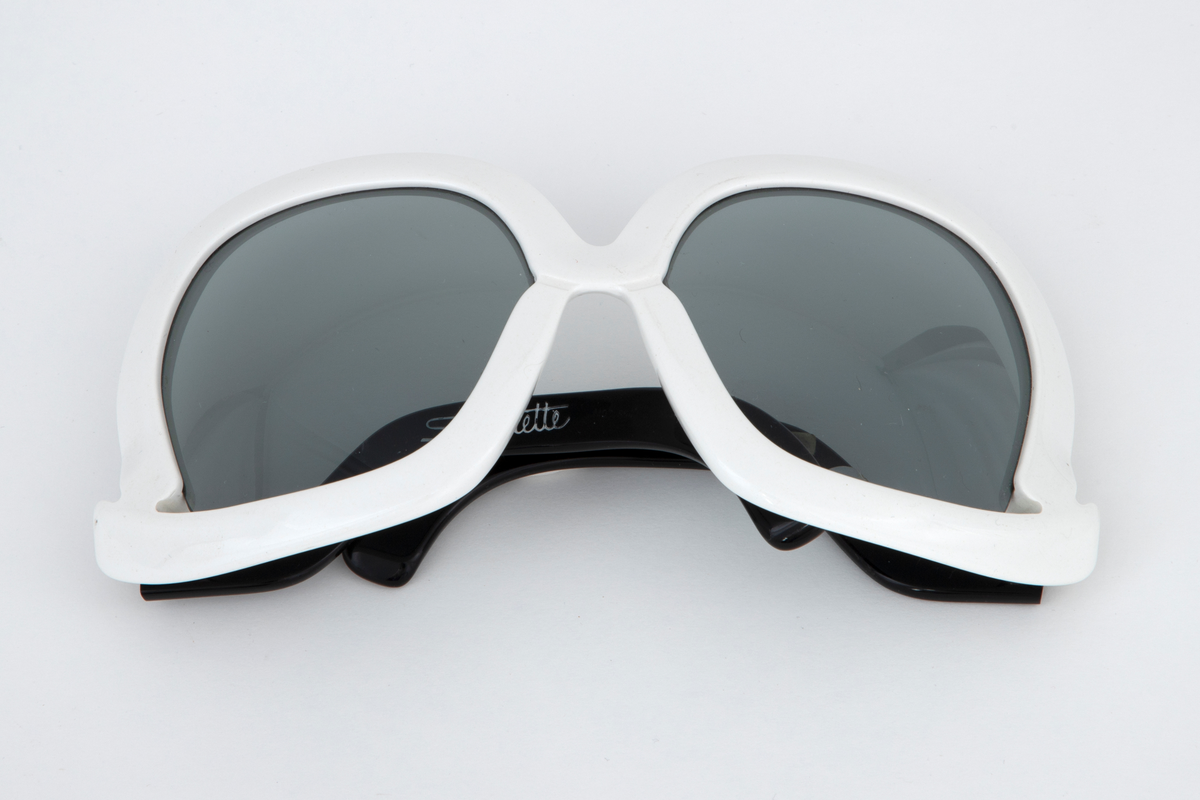 Store solbriller med hvit plastinnfatning og svarte, svungne stenger som er festet nederst på innfatningen. Grønne plastglass. Svart, rektangulært plastetui med åpning på høyre kortside.