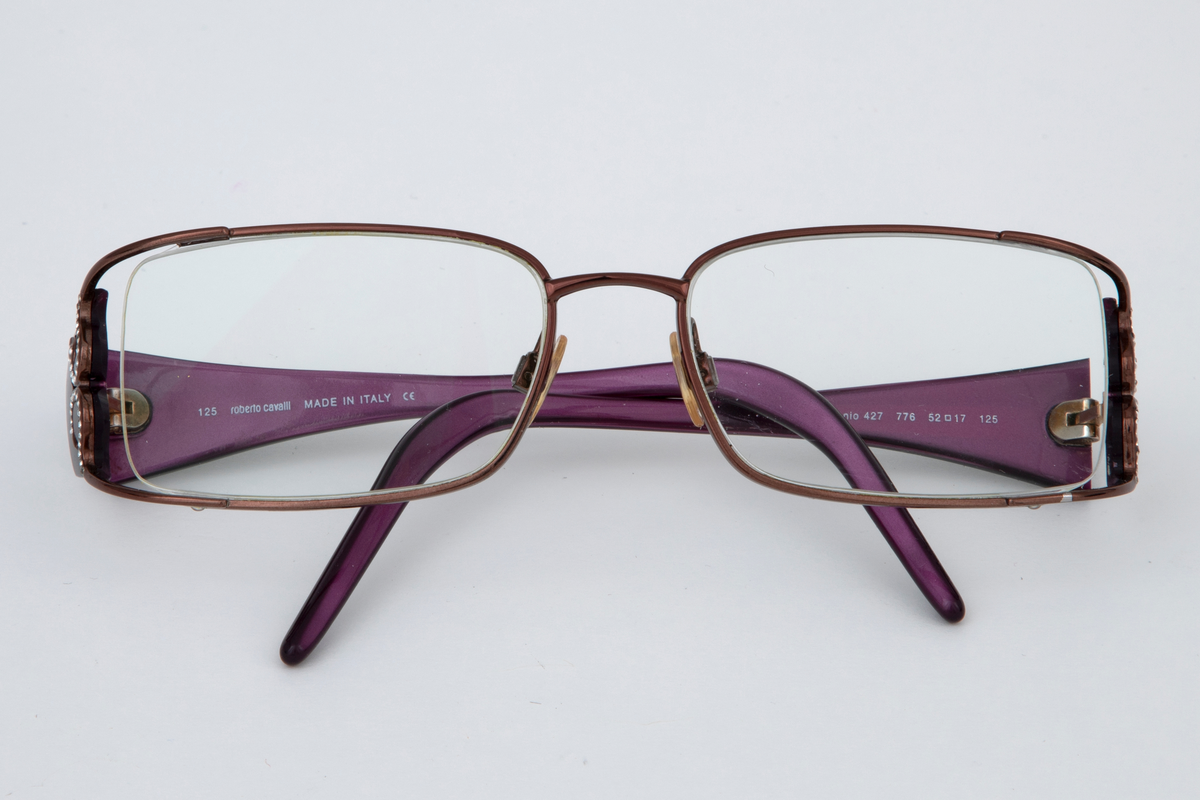 Damebriller med ovale, kantete glass i smal mørkerød metallramme og to slangeformede strassfigurer på hver side. Brede plaststenger som smalner mot de bøyde endene. På innsiden er stengene lilla. Svart brilleetui i hardplast med svart og hvitt dyretrykk på innsiden.