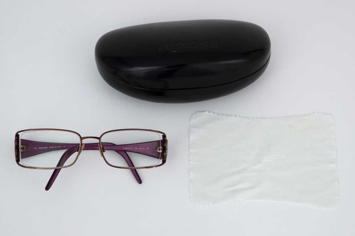 Damebriller med ovale, kantete glass i smal mørkerød metallramme og to slangeformede strassfigurer på hver side. Brede plaststenger som smalner mot de bøyde endene. På innsiden er stengene lilla. Svart brilleetui i hardplast med svart og hvitt dyretrykk på innsiden.