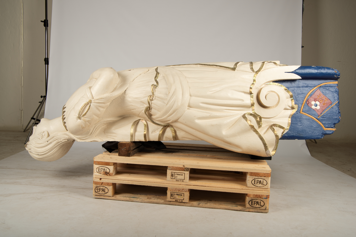 Helfigur, dame med gullrose i håret. Hvit kjole med gullkant, mørkt hår, blå sokkel med rødt emblem