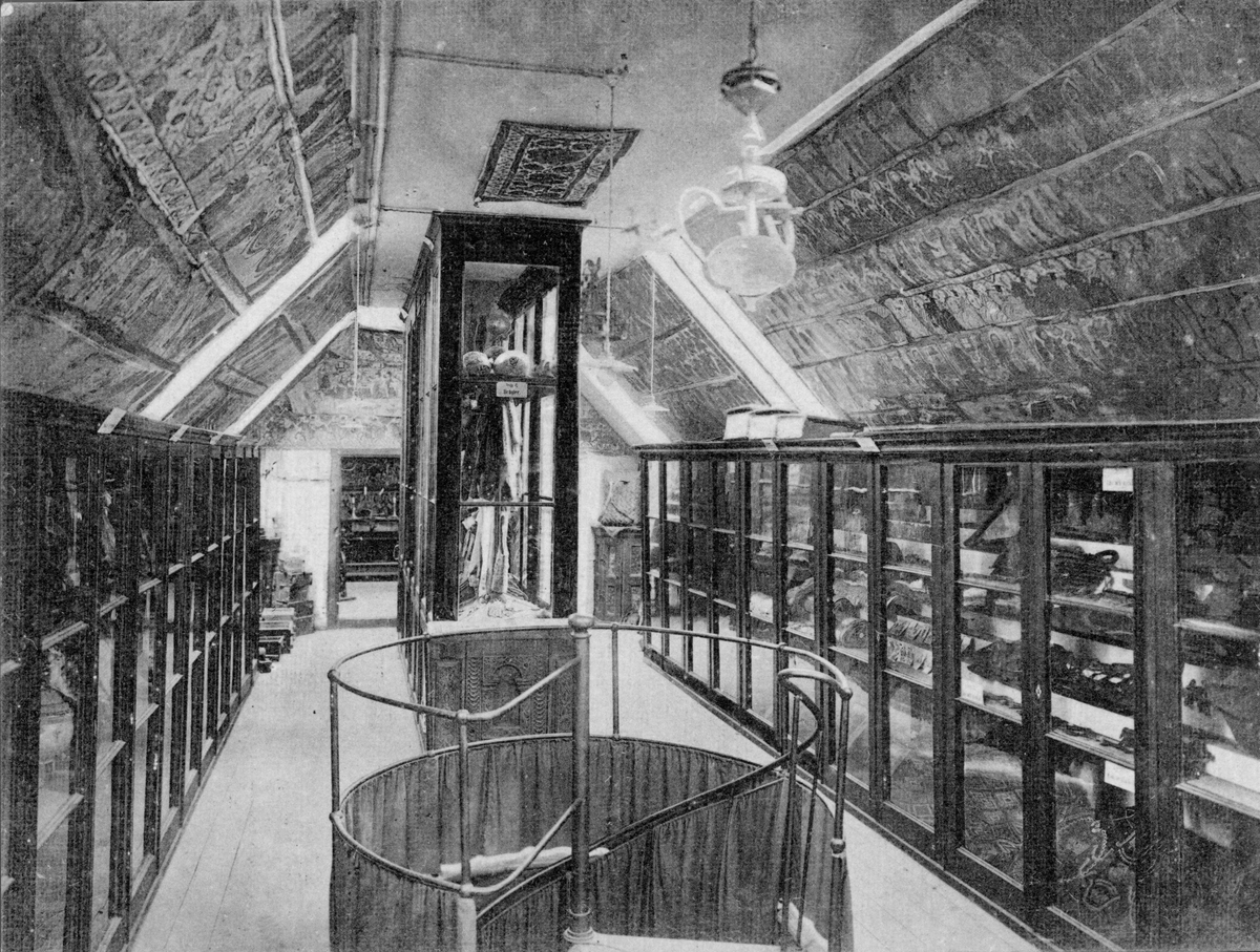 Vykort, "Hallands Museum, Halmstad." med noteringen "i Norreport". Interiör med föremål i montrar, målade bonader i taket och en spiraltrappa i förgrunden. Bild 2: Tonad variant i sin helhet.