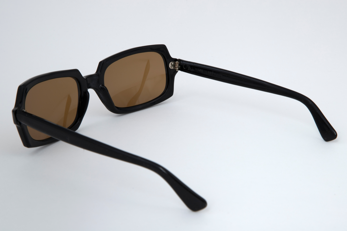 Solbriller med oval kantete plastglass og svart plastinnfatning med hengslede, svarte stenger.