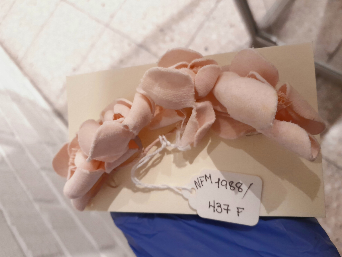 Seks ulike stoffblomar til pynt på drakt eller hatt. a: Bukett - orange/gul. b: Bukett - kvit/grøn. c: Rosett - svart/burgunder/rosa. d: Bukett - grøn/lilla. e: Bukett - grøn/lilla. f: grein - lys rosa. 