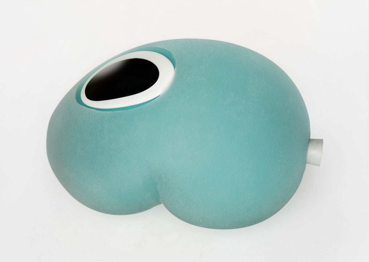 Ovalformet skulptur med organisk utforming, utført i polykromt overfangsglass. Skulpturens ytterside er dekket av klart glass som er mattet med sandblåsing, med unntak av et utspart felt på undersiden. Deretter lagvise sjikt i opak turkis, hvitt og sortfarget glass. På langsiden er et ovalt parti slipt vekk, slik at de indre sjiktene kommer til syne i form av konsentriske sirkler. På den ene kortsiden et hull med en gråfarget gummipropp.