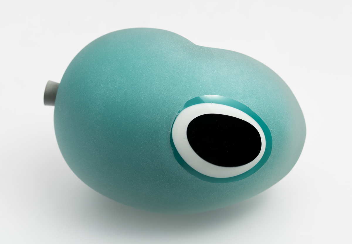Ovalformet skulptur med organisk utforming, utført i polykromt overfangsglass. Skulpturens ytterside er dekket av klart glass som er mattet med sandblåsing, med unntak av et utspart felt på undersiden. Deretter lagvise sjikt i opak turkis, hvitt og sortfarget glass. På langsiden er et ovalt parti slipt vekk, slik at de indre sjiktene kommer til syne i form av konsentriske sirkler. På den ene kortsiden et hull med en gråfarget gummipropp.