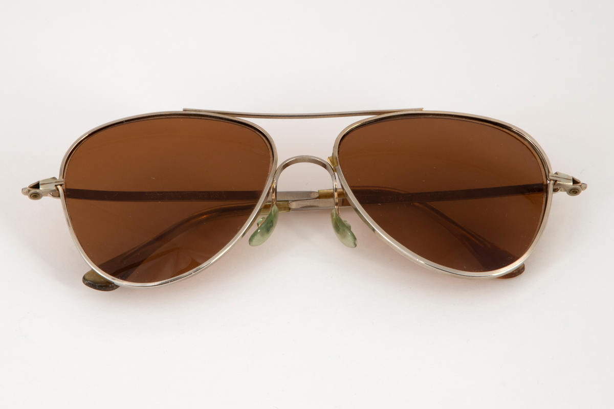 Solbriller med dråpeformede glass og smal metallinnfatning med en stang øverst, som forbinder glassinnfatningene. Smale, hengslede metallstenger som er plastbesatt på endene. Bevegelige neseputer i plast.